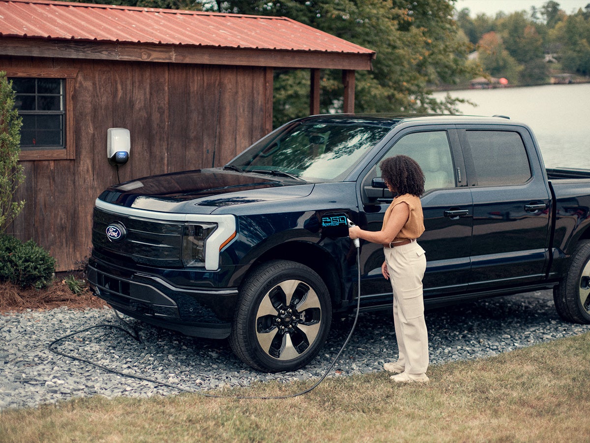 A woman charging her Ford F-150 Lightning using the Ford Connected Charge Station attached to her cabin in the background.